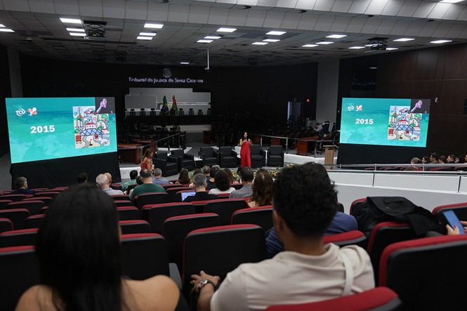 Foto horizontal: auditório do TJSC com público assistindo apresentação; telões exibem slide; ambiente institucional; tons preto, vermelho e verde.