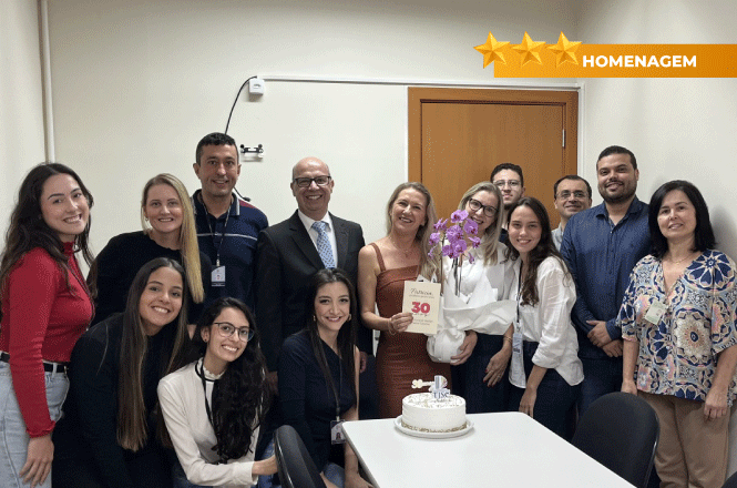 Foto horizontal: equipe celebra homenagem com bolo e flores; grupo sorridente em sala; clima festivo; tons branco, bege e azul.