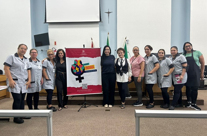 Foto horizontal: grupo de mulheres posa com banner do Programa Indira em sala institucional; ambiente formal