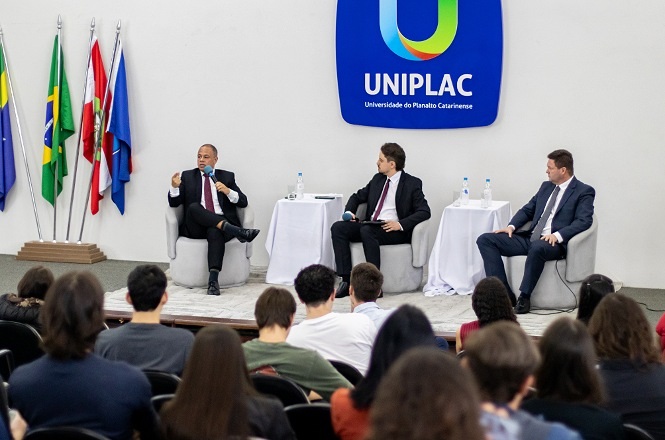 Foto horizontal: painel com autoridades em auditório da UNIPLAC; público assiste; bandeiras ao lado