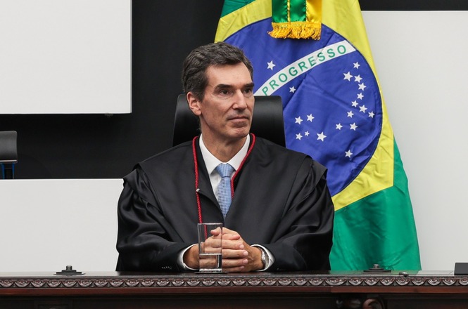 Foto horizontal: O desembargador Andreas Eisele em toga sentado em tribunal; bandeira do Brasil ao fundo; ambiente formal
