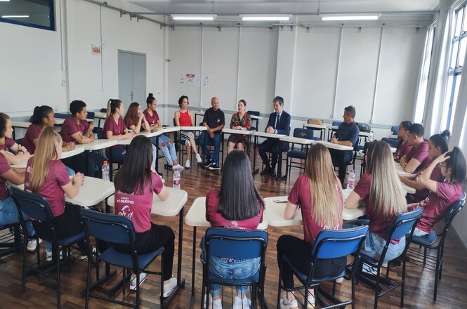 Foto horizontal: grupo de mulheres em roda, participa de encontro educativo; pessoas sentadas em sala; ambiente claro