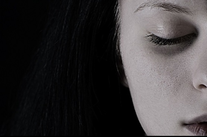 Foto horizontal: close de rosto feminino com olhos fechados; expressão introspectiva; fundo escuro
