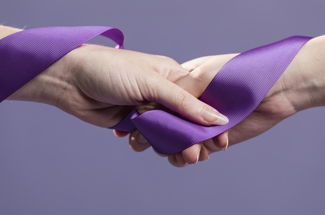 Foto horizontal: mãos entrelaçadas com laço roxo, símbolo de apoio e conscientização; fundo neutro