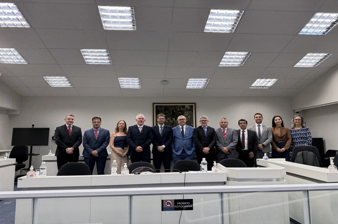 Foto horizontal: grupo de autoridades posa em sala institucional; alinhados atrás de bancada; ambiente formal