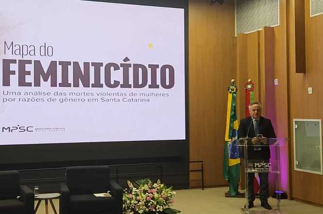 Foto horizontal: autoridade apresenta “Mapa do Feminicídio” em auditório; bandeiras ao lado; telão ao fundo