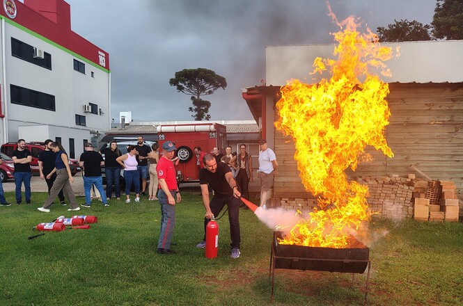 Foto horizontal: homem usa extintor para conter chamas altas; bombeiros e público observam; área externa