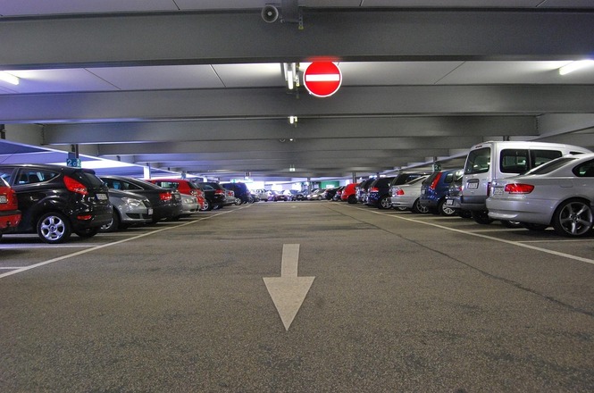 Foto horizontal: estacionamento coberto com carros alinhados; seta no chão indica direção; placa de proibido ao fundo