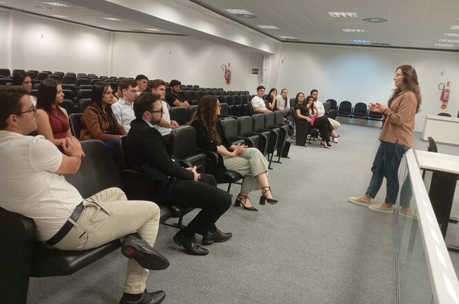 Foto horizontal: palestrante fala a grupo em auditório; público atento nas cadeiras; ambiente institucional