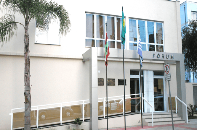 Foto horizontal: fachada de fórum da Comarca de Capinzal com bandeiras hasteadas e palmeira; entrada com rampa
