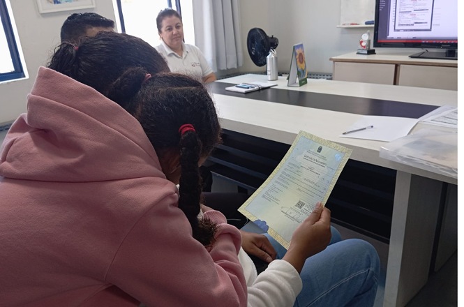 Foto horizontal: mãe com criança lê documento em atendimento; servidora ao fundo; ambiente de cartório