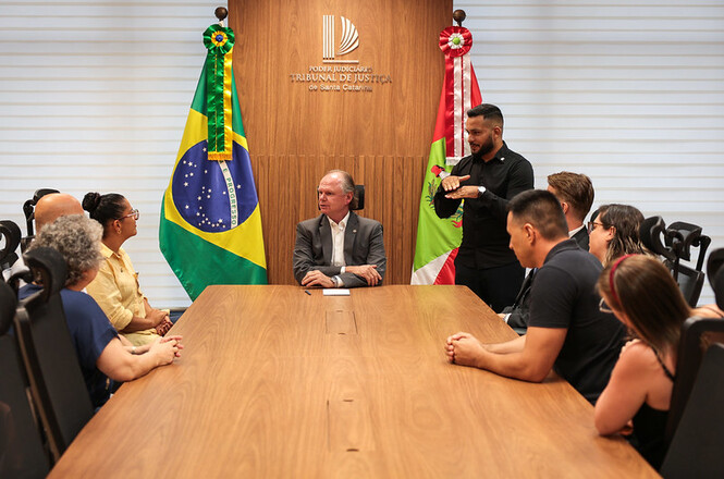 Foto horizontal: desembargador Rubens Schulz em reunião no TJSC com grupo e intérprete; tons de madeira, verde e amarelo.