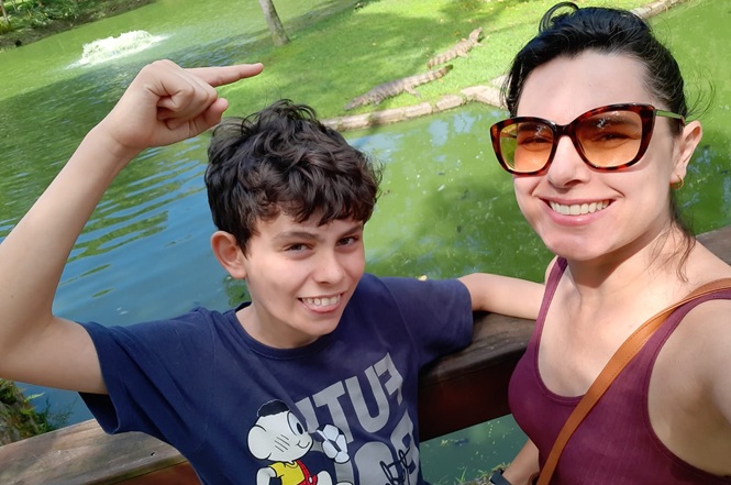 Foto colorida: Adriana Cechinel, servidora da Vara de Precatórios da comarca da Capital, com seu filho Giancarlo, de 12 anos, à beira de lago verde, paisagem natural externa, passeio ao ar livre, posando e apontando cenário em verde, azul e marrom