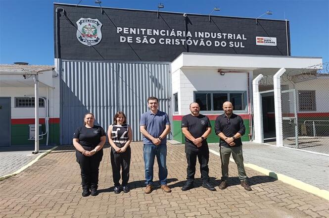 Foto colorida: grupo alinhado em frente à Penitenciária Industrial de São Cristóvão do Sul, área externa urbana, visita institucional, registrando encontro