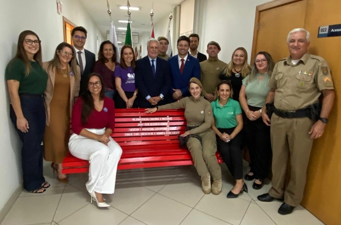 Foto colorida: grupo reunido em corredor institucional, campanha Banco Vermelho, ação de conscientização, posando junto ao banco em vermelho