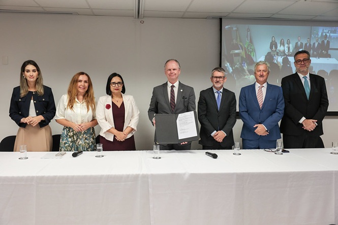 Termo assinado nas mãos do presidente do TJSC, desembargador Rubens Schulz, ao centro, com  grupo de autoridades, homens e mulheres em trajes formais, mesa solene, auditório interno, acordo institucional