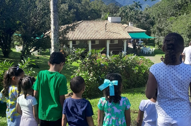 Foto colorida: crianças observando casa, paisagem verde rural, convivência educativa, admirando cenário em verde e marrom