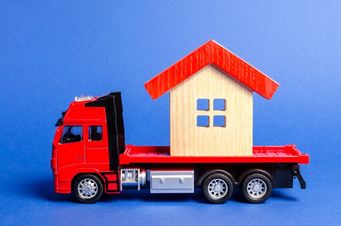Foto colorida: caminhão vermelho com casa de madeira, fundo azul neutro, transporte residencial, levando carga em vermelho, bege e azul