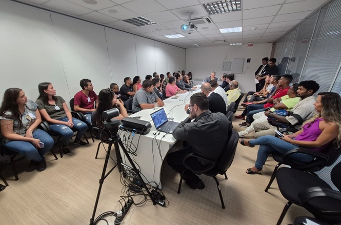 Foto colorida: comissão reunida em mesa longa, sala de reuniões, encontro institucional, debatendo pautas em branco, cinza e preto