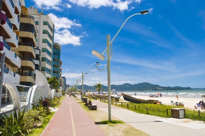 Foto colorida: orla urbana com calçadão e prédios, paisagem litorânea, lazer público, pessoas caminhando em azul, branco e verde
