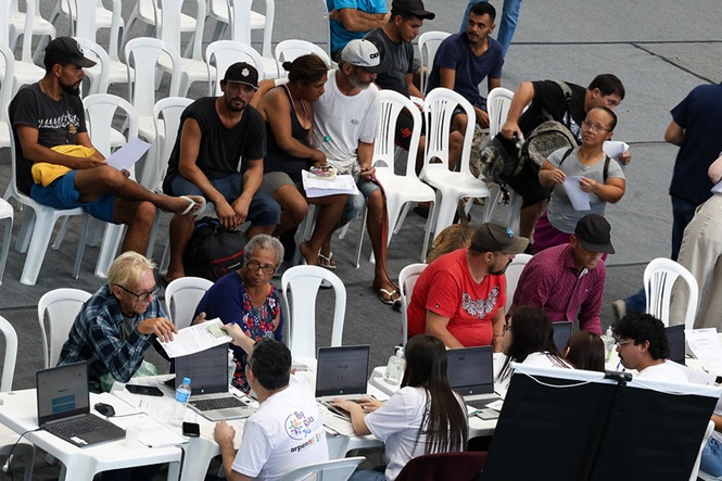 Foto colorida: atendimento em mesas do Centro Pop, ginásio interno do IEE, ação social, orientando público em azul, branco e cinza