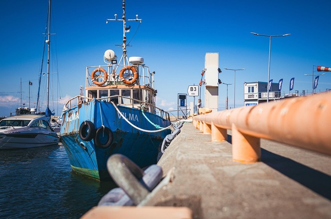Foto colorida: barco atracado em píer, marina urbana, logística portuária, aguardando operação em azul, laranja e cinza