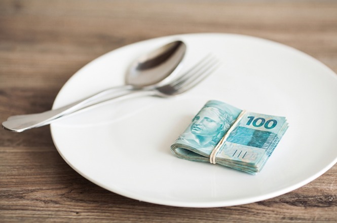 A imagem exibe notas de cem reais sobre um prato branco, com um garfo e uma colher ao lado.