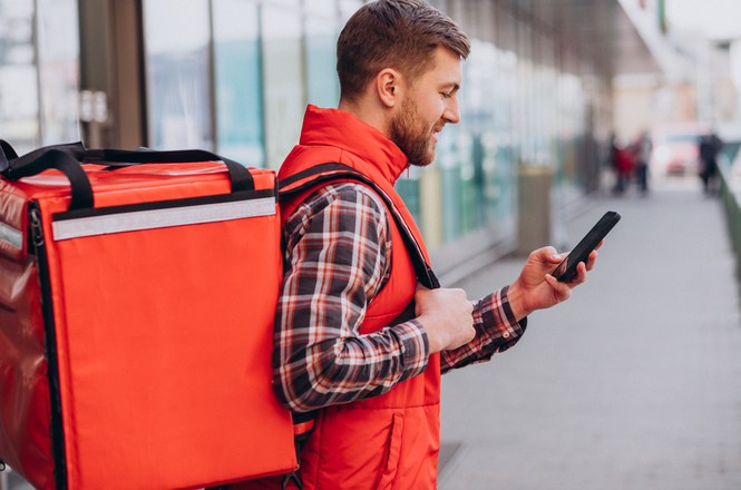 Entregador com mochila térmica, área urbana, trabalho por app, conferindo celular em vermelho e cinza