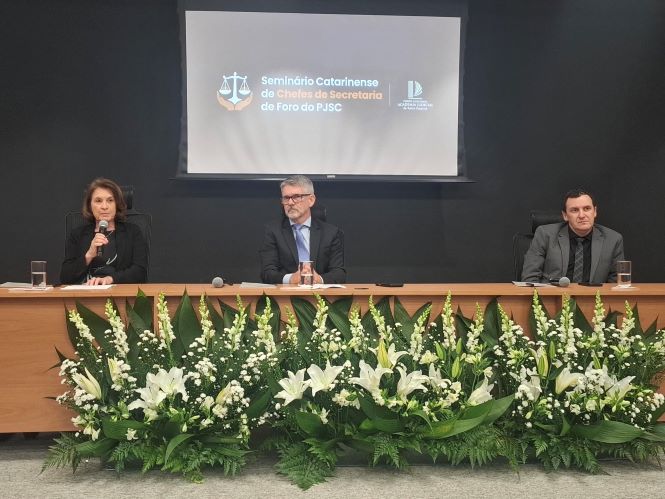 Em um auditório, três autoridades estão sentadas atrás de uma mesa de madeira, posicionadas lado a lado. À esquerda, a desembargadora Vera Lúcia Copetti segura um microfone e fala, com expressão atenta. No centro, o corregedor Dinart Machado está sentado, de frente, com postura formal. À direita, o diretor-geral administrativo Alexsandro Postali também está sentado, olhando em direção ao público. À frente da mesa, há uma longa decoração com flores brancas e folhagens verdes. Ao fundo, um telão exibe o nome do evento “Seminário Catarinense de Chefes de Secretaria de Foro do PJSC”, em ambiente institucional e sóbrio.