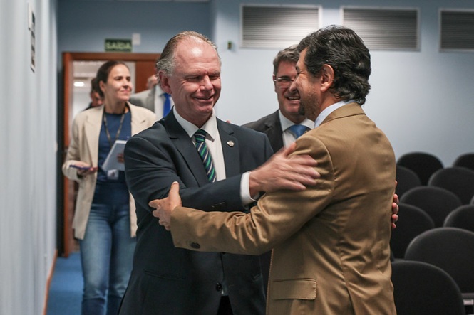 Dois homens de terno estão se abraçando calorosamente em um corredor, com outras pessoas sorrindo ao fundo.