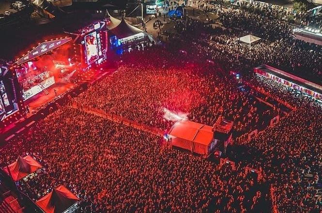 Uma vista aérea de um grande concerto noturno com uma multidão de fãs iluminada por luzes vermelhas no palco e efeitos de fumaça.