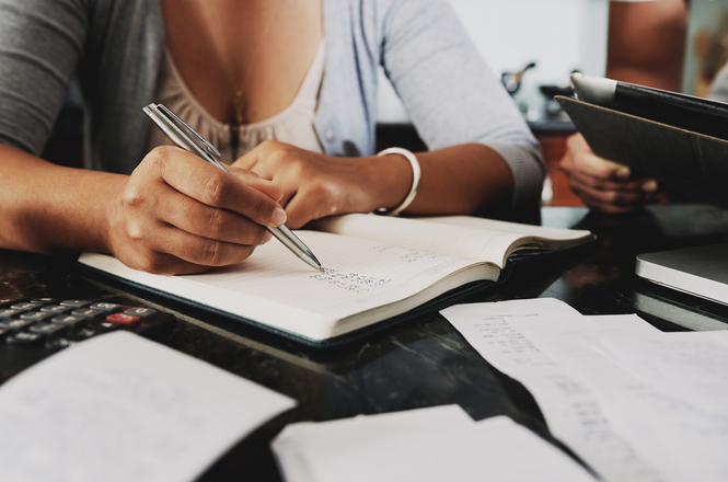 As mãos de uma pessoa segurando uma caneta, escrevendo em um caderno aberto, com recibos e uma calculadora sobre a mesa.