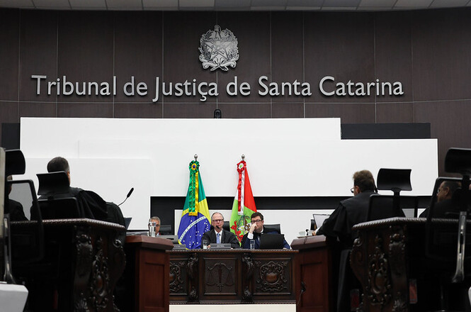 Uma cena formal de tribunal dentro do Tribunal de Justiça de Santa Catarina, com juízes e profissionais jurídicos sentados em um banco de madeira sob bandeiras e emblemas oficiais.