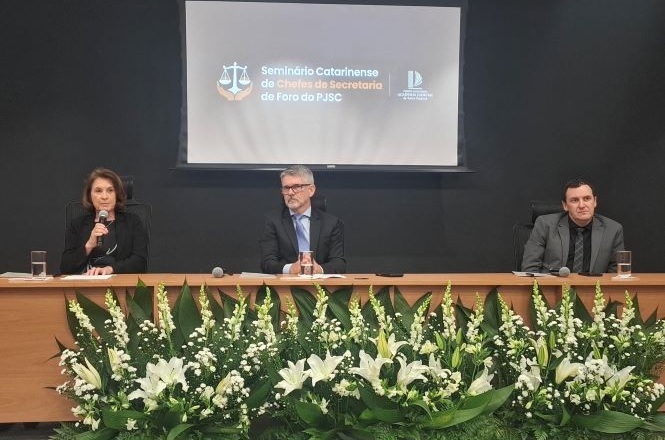 Três profissionais estão sentados em uma mesa de conferência durante um seminário formal, com uma tela grande exibindo os detalhes do evento atrás deles.