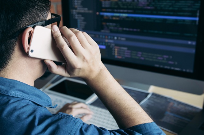 Uma pessoa usando óculos está falando ao telefone enquanto trabalha em um computador com código exibido na tela.