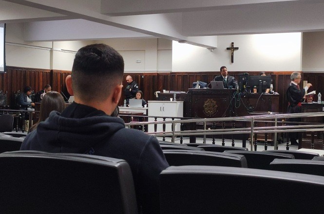 Uma cena de tribunal vista da perspectiva do público, mostrando pessoas sentadas e em pé enquanto um procedimento legal acontece.