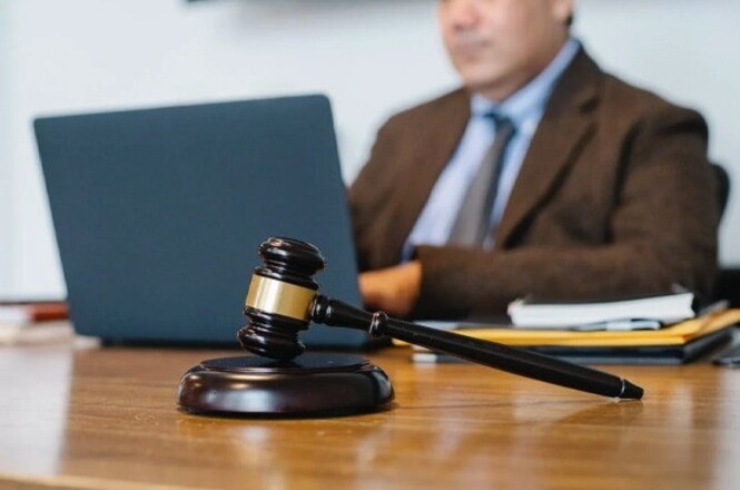 Um marreta de madeira repousa proeminentemente sobre uma mesa, com um advogado desfocado sentado atrás de um laptop no fundo, sugerindo um ambiente jurídico ou de tribunal.