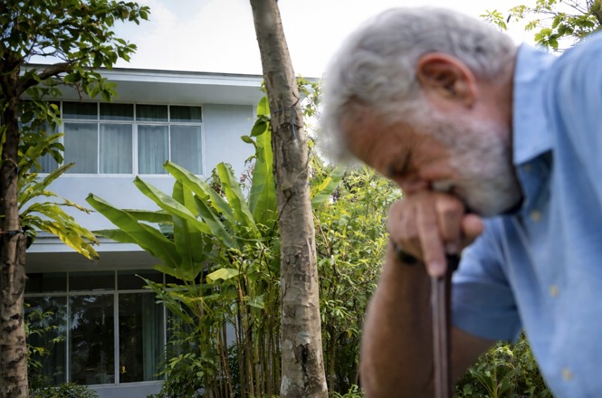 A imagem retrata um homem idoso, com barba grisalha e camisa azul clara, em um momento de aparente reflexão ou cansaço, apoiado em uma bengala.