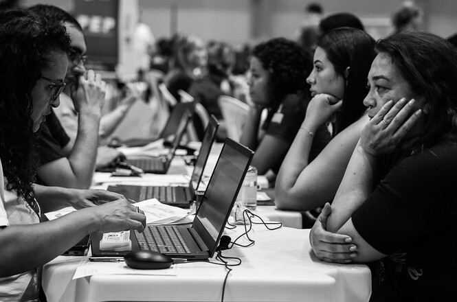 Foto em preto e branco pessoas em mesas com laptops, salão interno, atendimento, preenchendo dados em preto e branco