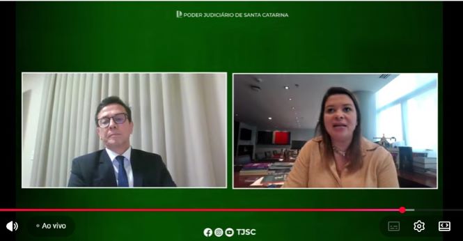 print de tela de uma transmissão ao vivo do Poder Judiciário de Santa Catarina, com fundo verde institucional e dois participantes em janelas lado a lado. À esquerda, está o diretor executivo da Academia Judicial, desembargador Edir Josias Silveira Beck, sentado diante de uma cortina clara. Ele veste terno escuro, camisa branca e gravata azul, usa óculos e aparece com expressão séria. À direita, está a diretora jurídica da Confederação Nacional das Seguradoras (CNSeg), Glauce Karine de Jesus Madureira Carvalhal, sentada em uma sala ampla e iluminada, com janelas ao fundo e mesa de reuniões ao lado. Ela veste blusa bege clara e colar discreto, com expressão atenta durante a fala. Na parte inferior da imagem aparecem os controles da transmissão e a indicação de que o evento está ao vivo.