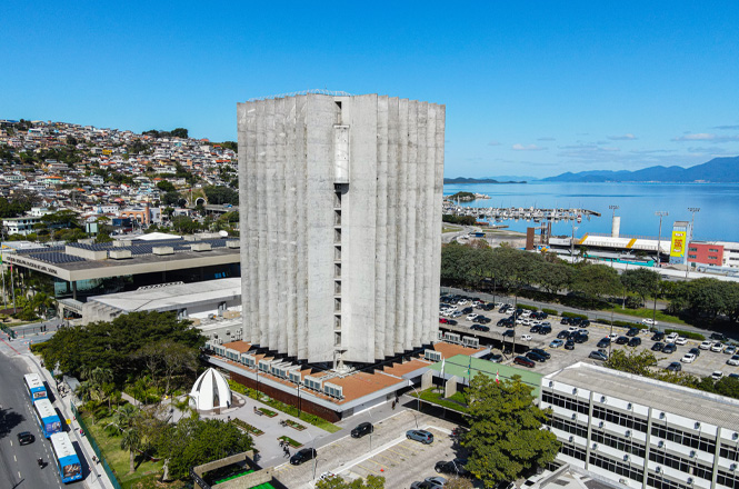 Foto colorida: edifício do TJSC, com o mar ao fundo, paisagem urbana costeira, serviço institucional