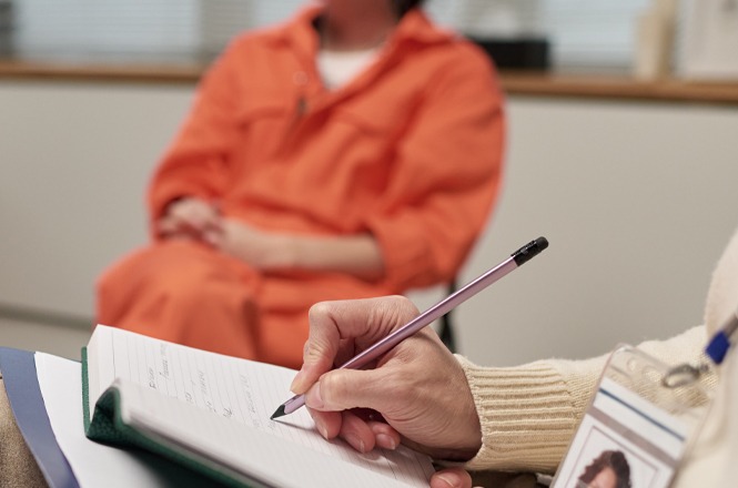 Foto colorida: pessoa vestida com traje laranja, sentada frente a profissional anotando, sala interna institucional, oitiva judicial, registrando depoimento