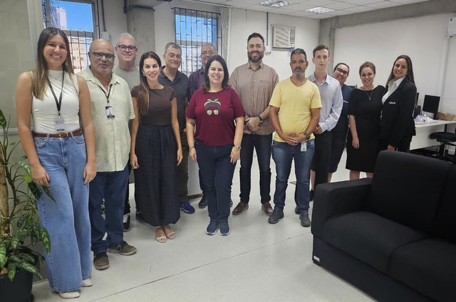 Foto colorida: grupo alinhado em sala administrativa, ambiente interno institucional, rotina de almoxarifado, posando para registro em branco, bege e preto