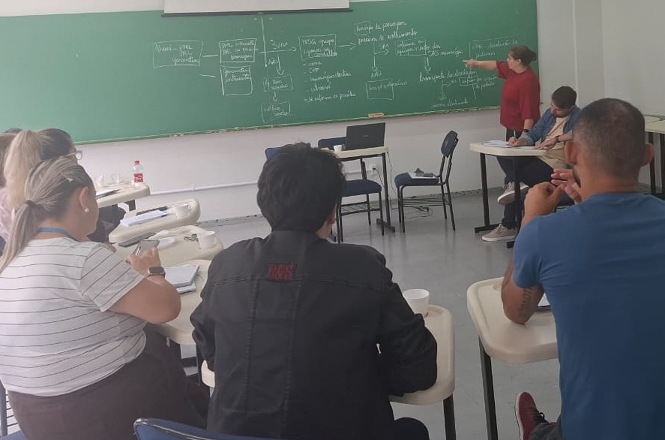 Foto colorida: pessoas sentadas em sala de aula, quadro verde ao fundo, reunião comunitária, debatendo temas em verde, branco e bege