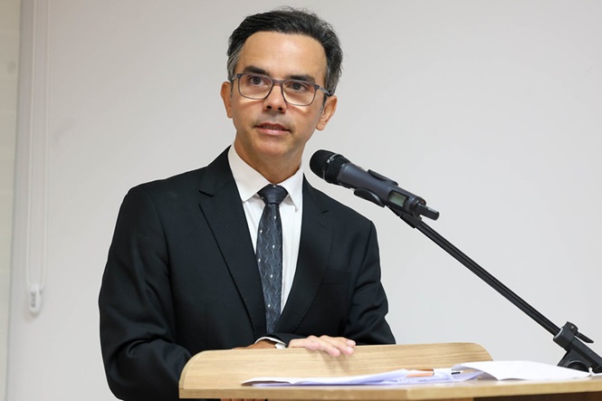 Fotografia horizontal: Humberto Goulart da Silveira está de terno escuro e fala ao microfone, fundo branco e púlpito de madeira em evento institucional