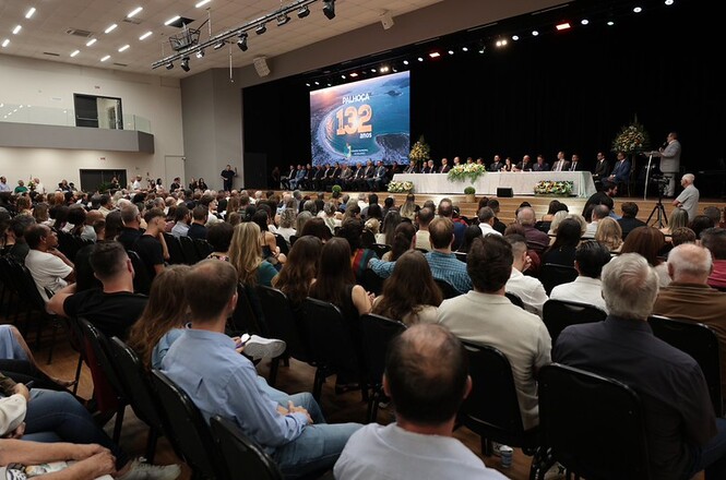 Fotografia horizontal: público e autoridades em auditório amplo com palco iluminado e telão azul em evento institucional, a plateia lotada assiste cerimônia, em tons preto, azul e branco