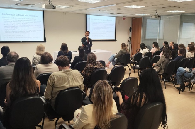 Fotografia horizontal: palestrante e público, em sala clara com telões e cadeiras pretas, evento institucional, homem fala ao microfone enquanto plateia observa, em tons branco, preto e bege