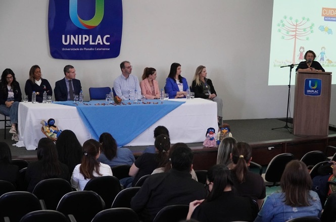 Fotografia horizontal: mesa de debatedores e público, auditório branco com logotipo azul da UNIPLAC,  palestrante fala ao microfone enquanto mesa acompanha, em tons azul, branco e preto