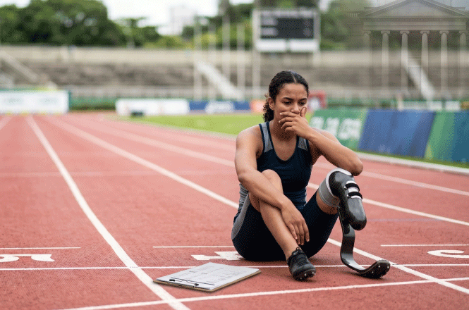 Foto colorida: atleta sentada em pista de corrida, estádio ao ar livre, esporte paralímpico, descansando após prova em vermelho, preto e verde