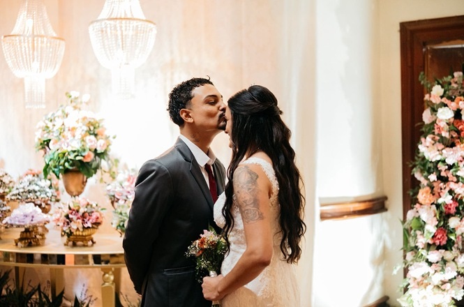 Foto colorida: casal em cerimônia de casamento comunitário, salão decorado com flores e lustres, união civil, celebrando momento em branco, dourado e rosa